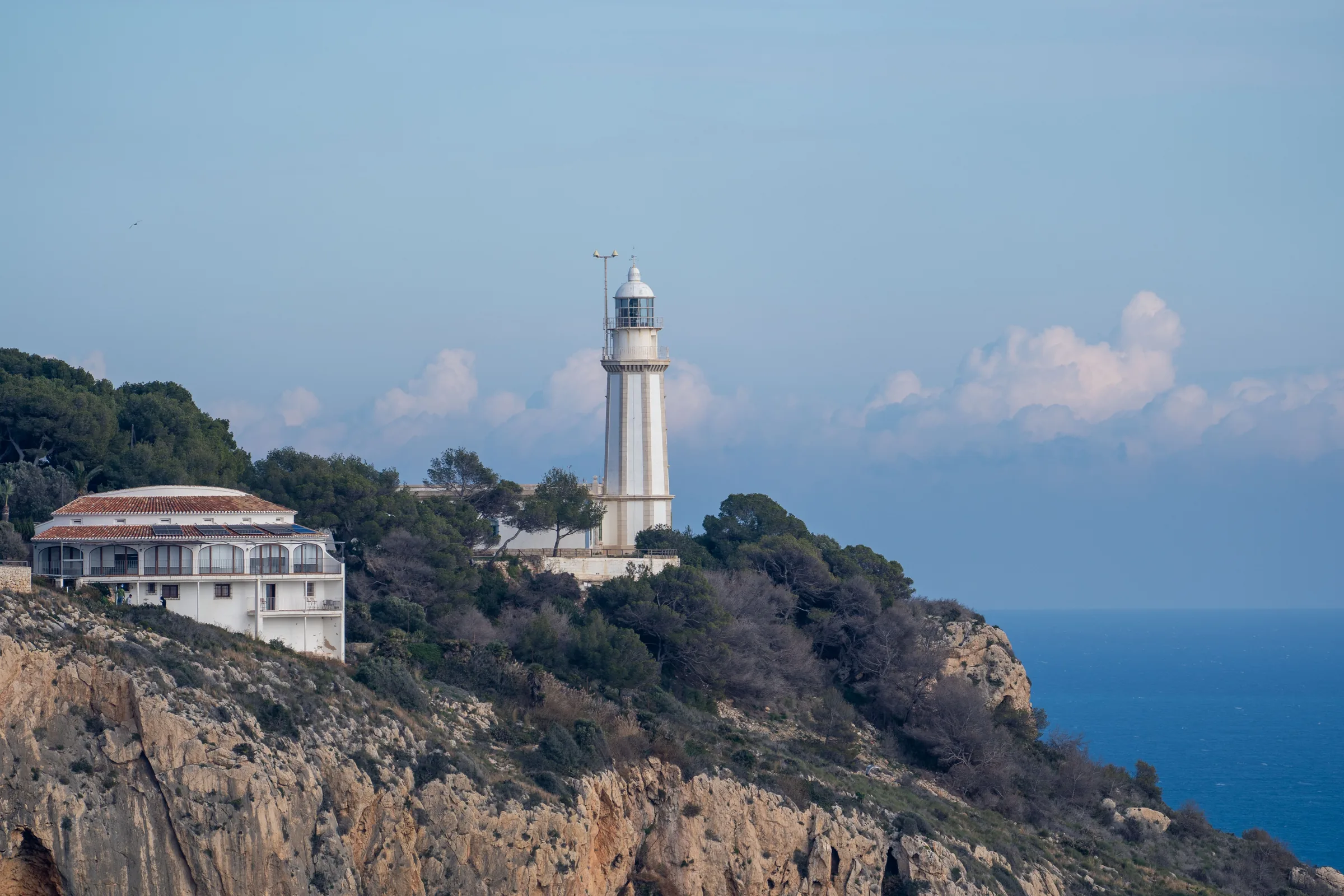 The white lighthouse of Cap de Sant Antoni among pines on the cliff, with the open sea behind