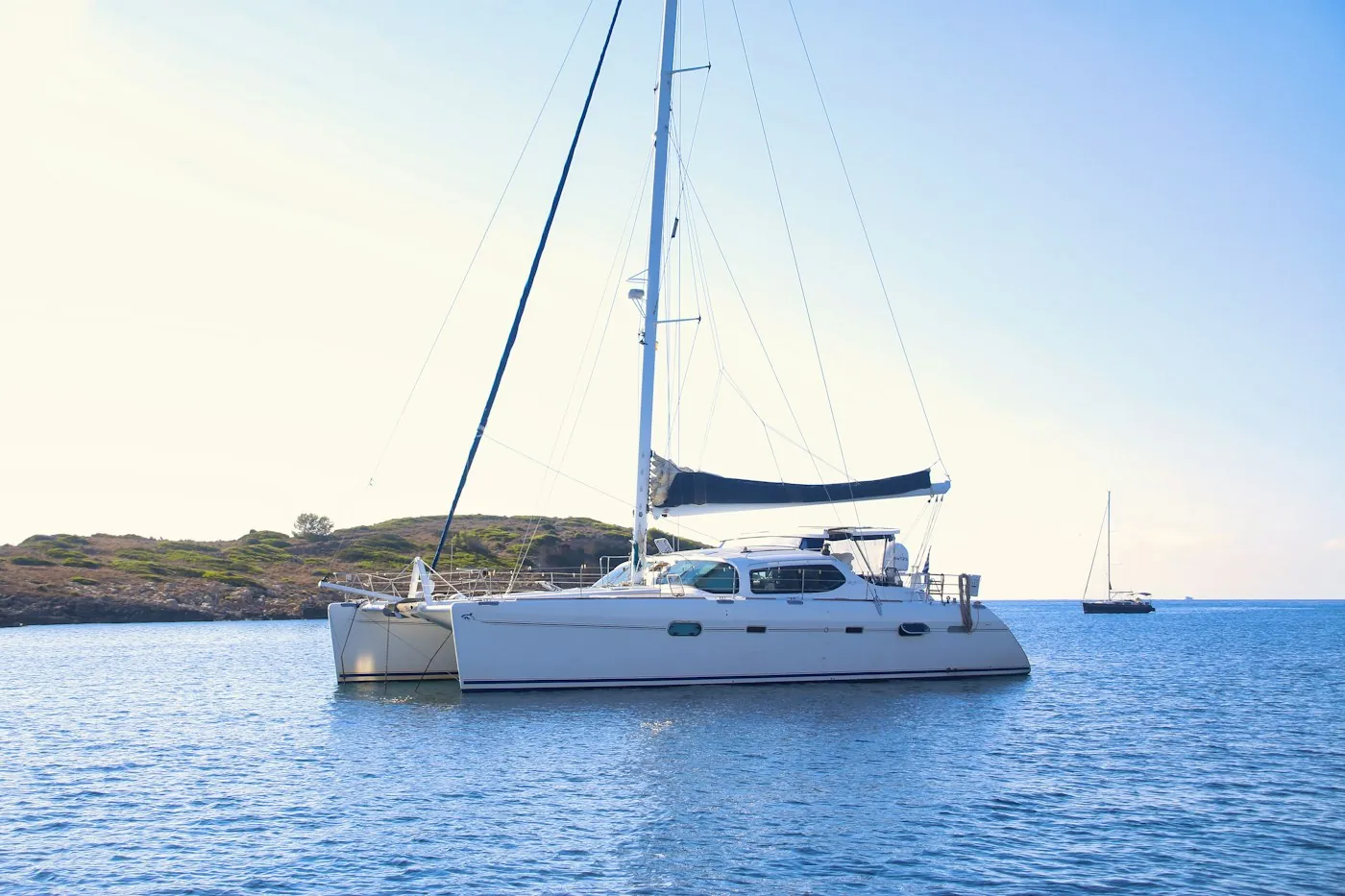 White sailing catamaran at anchor on calm water near a low headland